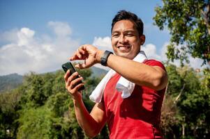 A happy Asian man in sportswear checks his calorie count on his smartwatch while jogging in a park. photo
