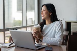 An Asian woman enjoying a hot drink at a coffee shop while taking a break from work on her laptop. photo