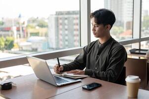 A focused businessman working in his office, writing in a book while sitting in front of a computer. photo