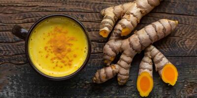 Cup of yellow liquid with frothy top on wooden table next to turmeric roots, gastronomy photography photo