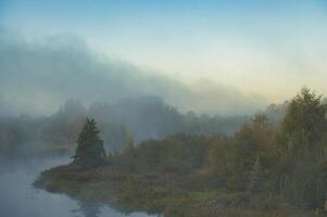 Misty morning landscape featuring a tranquil river with trees reflected in the calm water photo