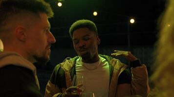 Friends enjoy a lively gathering under string lights while sipping drinks in a cozy outdoor setting at night video