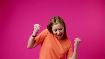A cheerful woman in a vibrant orange t-shirt enjoys dancing by herself, smiling and moving energetically to an unheard rhythm against a striking pink backdrop. video