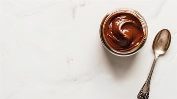 Clear jar of hazelnut spread on white background with metal spoon beside it, gastronomy photography photo
