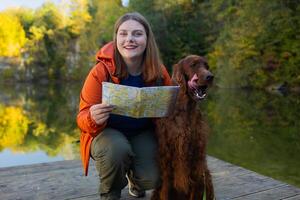 Young woman traveling with dog. Beautiful female sitting near a lake together with Irish Setter dog and looking around. Happy girl interact with her pet at hiking rest with mountain and forest view. photo