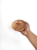 The image shows a hand holding a round wooden bowl with a smooth surface. The plain white background emphasizes the natural texture and grain patterns of the wood, making the bowl the central focus. photo