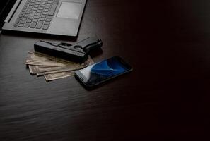 Computers, mobile phone, money, guns, and other devices that hackers use to steal or hijack information to trick victims into taking money. laying on wooden table in dark room in mysterious apartment. photo