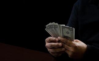 Hands of a man wearing a black shirt, sitting on a chair and a table, is a thief, holding money, counting the amount obtained from hijacking or robbing, in a dark background photo