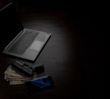Computers, mobile phone, money, guns, and other devices that hackers use to steal or hijack information to trick victims into taking money. laying on wooden table in dark room in mysterious apartment. photo