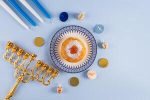 Fried donut with powdered sugar and jam on a plate, candles, minor, dreidels, gold coins on a blue background. The concept of the Hanukkah. photo
