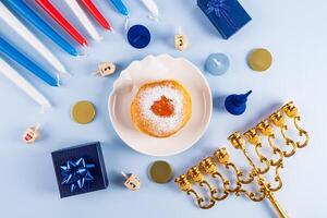 Fried donut with powdered sugar and jam on a plate, candles, minor, dreidels, gold coins on a blue background. The concept of the Hanukkah. photo
