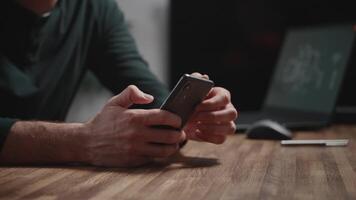 Close-up of male hands using a modern smartphone. A man is sitting at a table and using online apps. A laptop is in the background. video