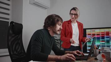 Colleagues working in a design and architecture studio. A woman interior designer in a red jacket and glasses is discussing a project with a male architect. The workspace features palettes video