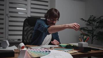 A stylish male architect is working on creating a project. The specialist is sitting at his workspace designing the interior. The worker is using blueprints, palettes, material samples, and textures. video