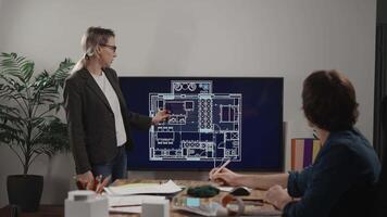 A woman architect is presenting a project in a design studio office. The plan of an apartment is on the screen, and a colleague is giving comments about the blueprint. video
