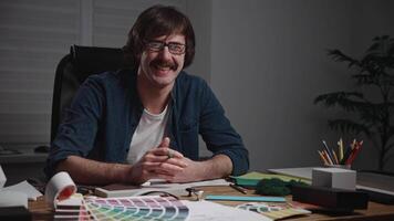 A portrait of an attractive man with a mustache. A successful architect-designer is at his workstation, smiling and looking at the camera. Bright studio with a wooden desk and designer tools. video
