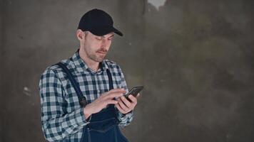 A man in overalls with a tartan cap is using a cell phone video