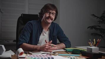 Portrait of a stylish man in glasses and with a mustache. The architect designer is at his workspace. On the wooden table lie palettes, textures, material samples, and blueprints. A positive hipster video