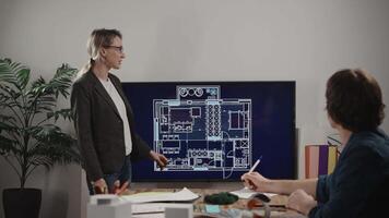A woman is giving a presentation of an architectural project. On the large screen, there is a house plan with furniture layout. A male colleague is listening to the designer and commenting video
