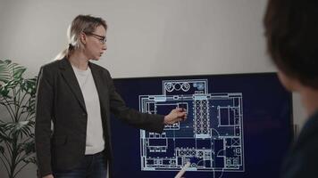 A woman architect is presenting a project in a design studio office. The plan of an apartment is on the screen, and a colleague is giving comments about the blueprint. video