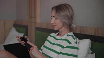 Women use a smartphone to communicate and work. Blonde woman on a bed in a hotel room, typing messages on her phone, and working in apps. video