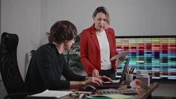 Colleagues working in a design and architecture studio. A woman interior designer in a red jacket and glasses is discussing a project with a male architect. The workspace features palettes video