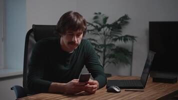 A stylish man with a mustache sits at a table using a smartphone. A modern laptop is on the table, and a blueprint or apartment plan is displayed on the screen behind. video