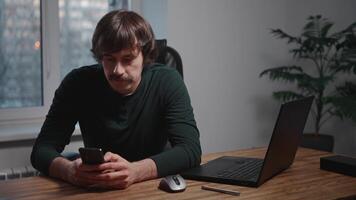 An attractive and stylish young man is sitting at his workspace using a smartphone. The hipster with a mustache is using mobile apps and browsing the mobile internet. Wooden table and a laptop video