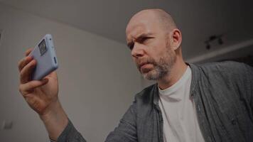 A man with a thoughtful expression examines his phone closely, holding it up in one hand, conveying curiosity or confusion. The neutral background emphasizes his focus on the device. video