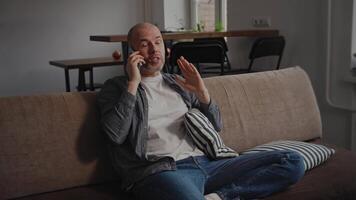 uma homem senta confortavelmente em uma sofá, falando em a telefone com uma alegre expressão, gesticulando com dele mão. a acolhedor interior características uma cozinha mesa e cadeiras dentro a fundo. video