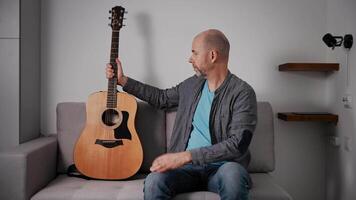 A middle-aged man with a short beard and shaved head is sitting on a gray couch, holding an acoustic guitar. The man is examining the guitar closely, possibly tuning it or inspecting its build. video