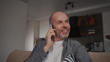 A man sits comfortably on a sofa, talking on the phone with a cheerful expression, gesturing with his hand. The cozy interior features a kitchen table and chairs in the background. video