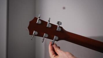Close-up of a hand adjusting the tuning peg on an acoustic guitar headstock. The wooden neck and metal tuners are in sharp focus, highlighting the precision in tuning the instrument. video