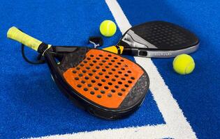 A padel or paddle tennis racket and a bright yellow tennis ball lie on a blue court. The net and a blurred figure of a player are visible in the background. photo