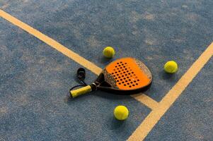 A padel or paddle tennis racket and a bright yellow tennis ball lie on a blue court. The net and a blurred figure of a player are visible in the background. photo
