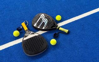 A padel or paddle tennis racket and a bright yellow tennis ball lie on a blue court. The net and a blurred figure of a player are visible in the background. photo