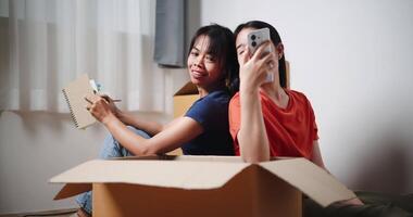 Handheld shot, Older sister is writing a shopping list in a notebook while the younger sister is shopping for household items via a mobile app. Moving to a new home concept video