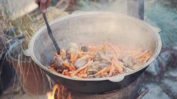 Chef mixes meat with vegetables and oil with slotted spoon in large cauldron. Appetizing dish of Oriental cuisine made of fried beef, carrots and onions in kazan. Tourist lunch at campfire. video