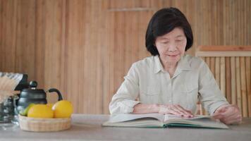 Senior asiatisch Frau Sitzung lesen ein Buch, Früchte und Teekanne auf das Tabelle im Bambus Haus, ruhig lesen ein Buch und genießen ein ruhig Lernen und entspannend Moment. Hobby, Selbstverbesserung Konzept. video