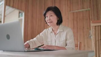 Elderly woman doing call using laptop on table in bamboo house, online learning, call with family, new way of life, searching information on internet, telecommunication concept. video