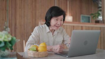 mayor mujer vocación utilizando ordenador portátil y hablando con contento cara en mesa en bambú casa.con familia, buscando información, mayor ciudadano estudiando en línea video