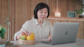 Senior woman doing l using laptop and picking up lemon and talking at table in bamboo house. with family, searching information, senior citizen studying online, online education concept. video