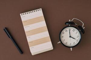 Planner pad, alarm clock and pen on brown background. Planner for organization and goal priorities. Writing down tasks and schedule reminders on office desk. Trendy brown color flat lay. photo