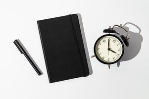 Black notebook, alarm clock and pen on white background. Planner for organization and goal priorities. Writing down tasks and schedule reminders on office desk. Minimal creative concept. Flat lay. photo