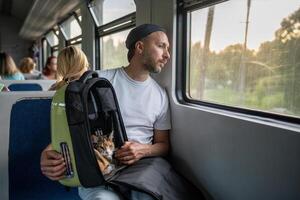 Serene man holds backpack with cat on lap sit in train car. Journey by railway transport with feline photo
