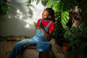 Relaxed black girl in earphones holds smartphone typing message, listen music in room with monstera. photo