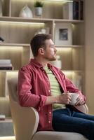 Thoughtful man slowly sips coffee while looks out window from armchair at home. Slow life, wellbeing photo