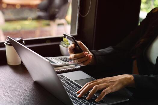 Business professional using smartphone and laptop while multitasking at a workspace by the window, focusing on digital communication and productivity during remote work in a modern office setting. photo
