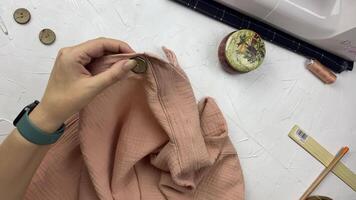 Women's hands create clothes from cotton muslin. Seamstress sews a wooden button to shirt using thread and needle. Hobbies and crafts. Workplace of a fashion designer on a white textured table. video