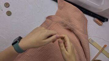 Seamstress sews a wooden button to shirt using thread and needle. Women's hands create clothes from cotton muslin. Hobbies and crafts. Workplace of a fashion designer on a white textured table. video